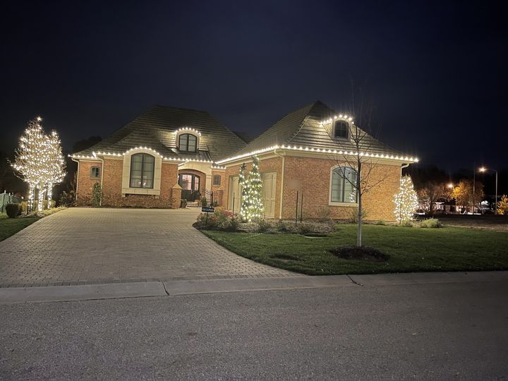 A brick French country home at night with its rooflines and dormers traced in warm white bulbs, and three trees wrapped trunk to tip in lights