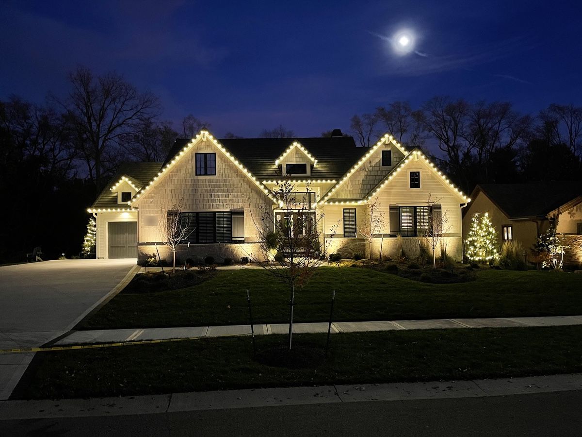 A white shingled two-story home at dusk under a full moon, every gable and roof edge outlined in warm white C9 bulbs, with a lit tree on each side of the lawn