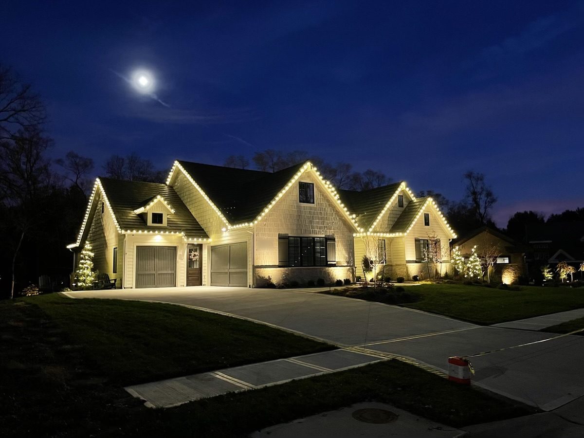 A cream shingled home with four gables outlined in warm white C9 bulbs under a full moon, with small lit trees by the garage and front beds